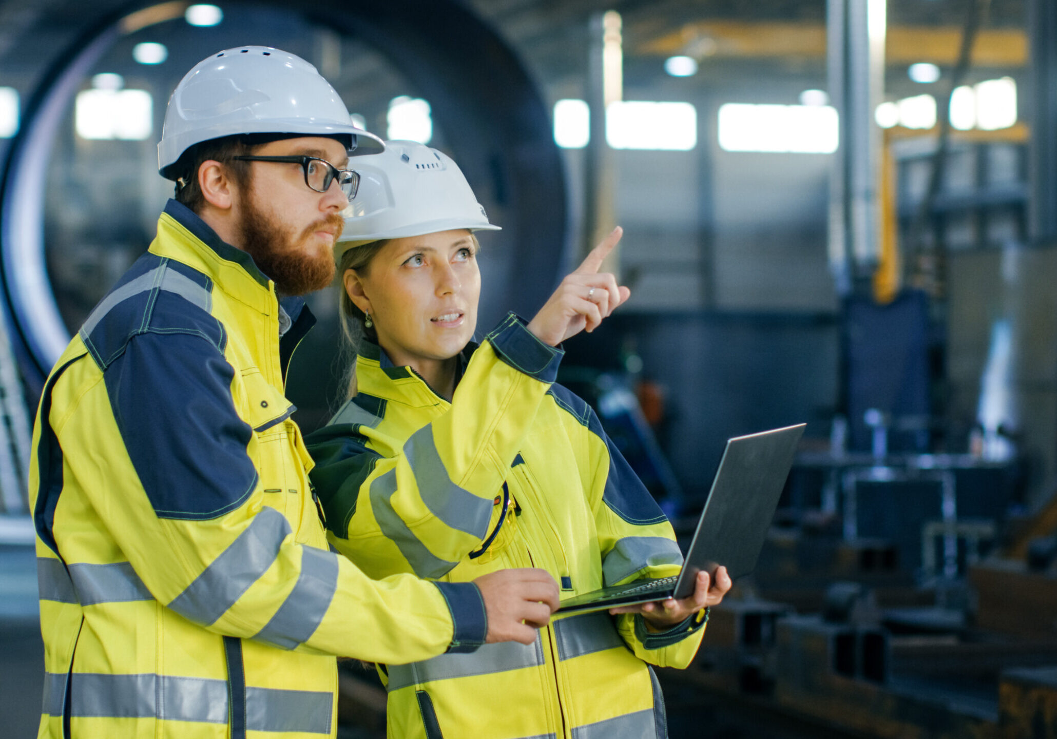 Portrait of Male and Female Industrial Engineers in Hard Hats Discuss New Project while Using Laptop. They Wear Safety Jackets.They Work at the Heavy Industry Manufacturing Factory.