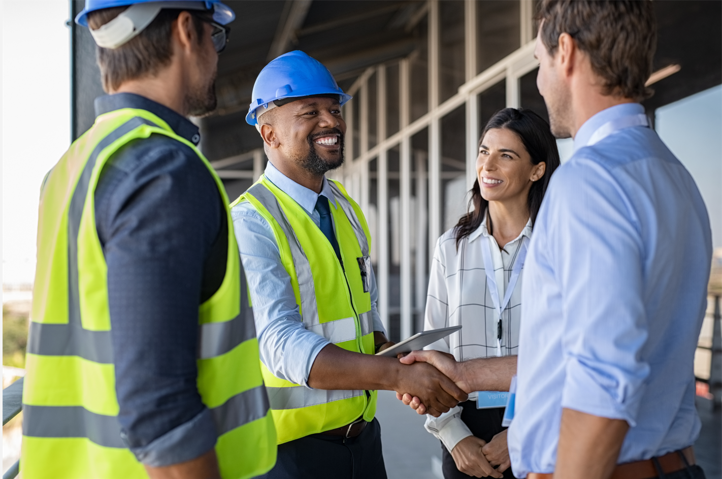 Four professionals on a construction site shake hands and smile, wearing safety helmets and high-visibility vests.